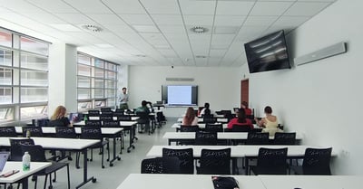 A classroom with rows of desks and chairs occupied by students facing a lecturer and a display. Two white Nureva HDL410 microphone and speaker bars are mounted on the front wall and a side wall.