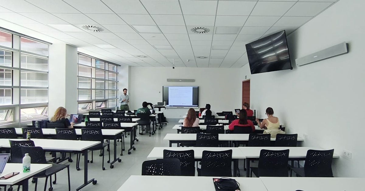 A classroom with rows of desks and chairs occupied by students facing a lecturer and a display. Two white Nureva HDL410 microphone and speaker bars are mounted on the front wall and a side wall.
