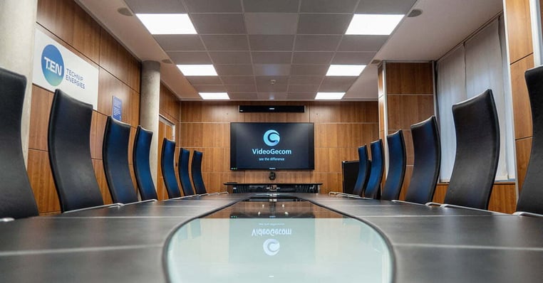 Executive boardroom with a long glass conference table, black high-back chairs and a wall-mounted display. Above the display is a Nureva HDL310 audio conferencing microphone and speaker bar for large hybrid meeting spaces.