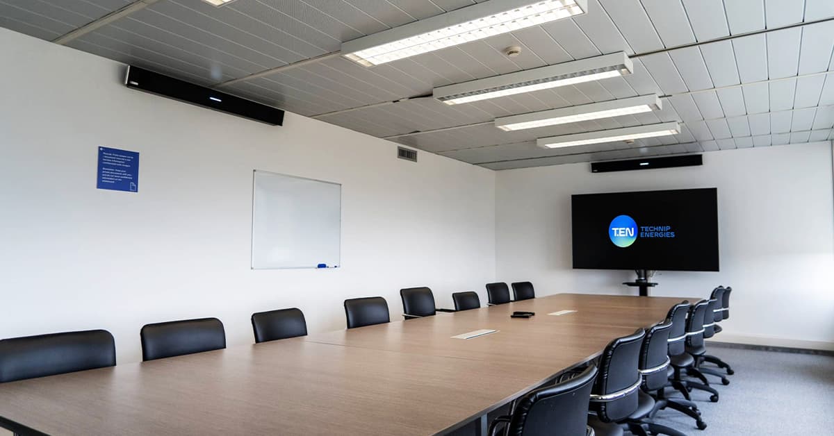 Large conference room with a long wooden table, black chairs, whiteboard and display. Mounted on the walls are two Nureva HDL410 audio conferencing microphone and speaker bars for clear audio in hybrid meetings.