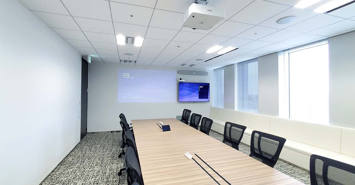 A long conference room with a central table and mesh-backed chairs, featuring a wall-mounted display and an off‑center white Nureva HDL310 audio system installed above it.