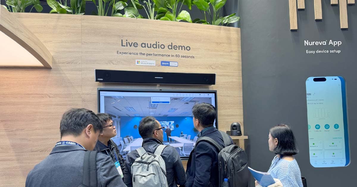 A group of attendees gets a live audio demo at the Nureva ISE 2026 stand, where a wall-mounted HDL audio system and a large screen display a remote meeting room. The backdrop features wood paneling, plant accents and signage promoting the Nureva App for device setup.