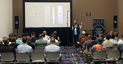 HDX full-featured audio demo in an InfoComm meeting room. A presenter is speaking to rows of seated attendees, alongside a display showing the HDX audio bar and actual working HDX components on stands.