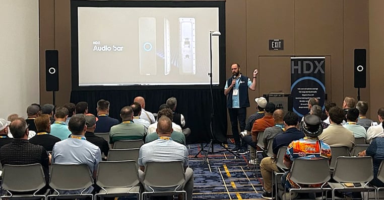 HDX full-featured audio demo in an InfoComm meeting room. A presenter is speaking to rows of seated attendees, alongside a display showing the HDX audio bar and actual working HDX components on stands.