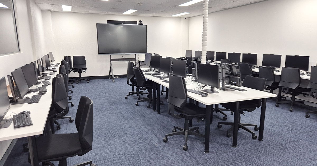 Canterbury Institute of Management classroom with rows of desks equipped with desktop monitors, keyboards and chairs. A large interactive display is positioned at the front of the room with a Nureva HDL200 system and video conferencing camera mounted on it..