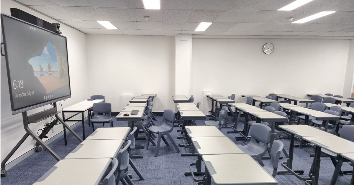 Canterbury Institute of Management classroom with multiple rows of gray desks and chairs. A large interactive display with a Nureva HDL200 audio system is mounted on a stand at the front of the room.