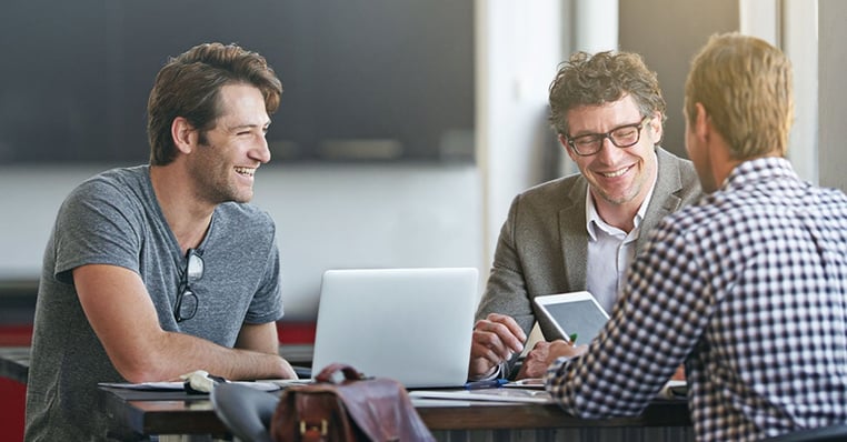 Three IT professionals have a conversation in an office setting. One has a laptop and one has a tablet.