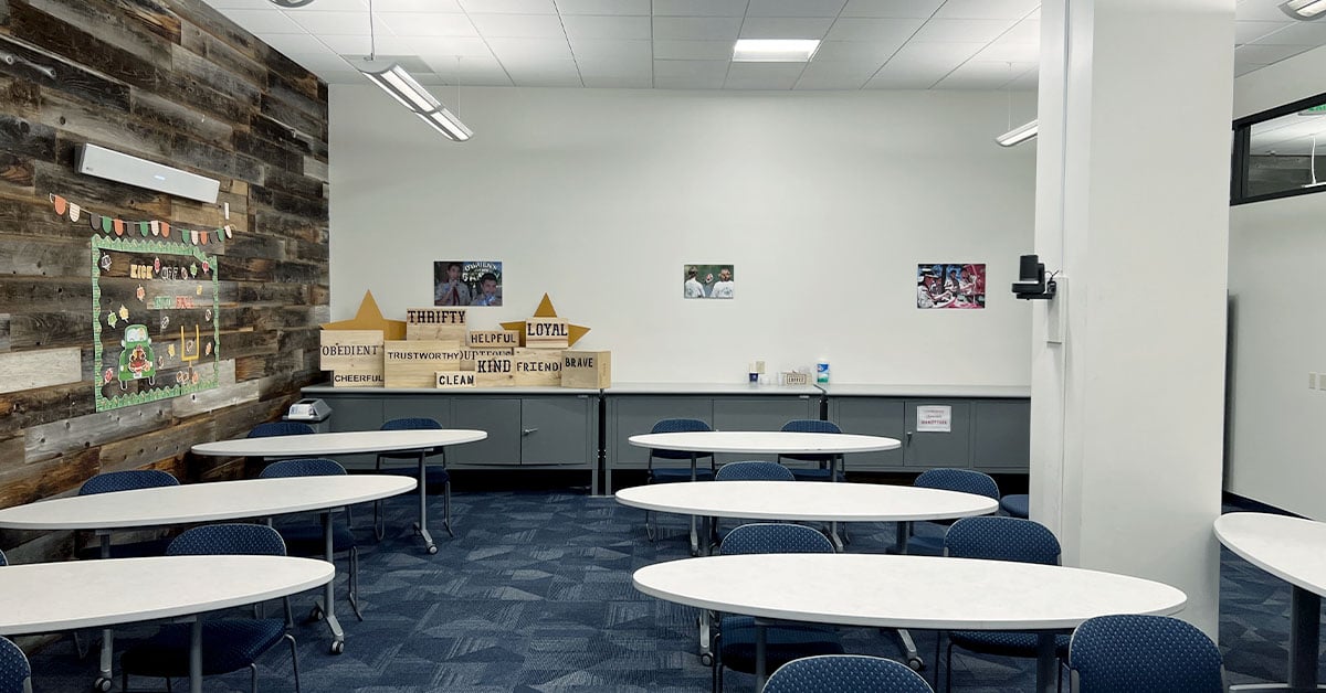 A multipurpose room with several tables and chairs, a large pillar, a back table and a wood accent wall with a Nureva microphone and speaker bar