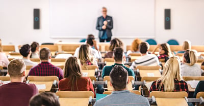 An instructor stands in front of a higher education classroom full of students. Two Nureva HDX audio bars are at the front of the classroom.