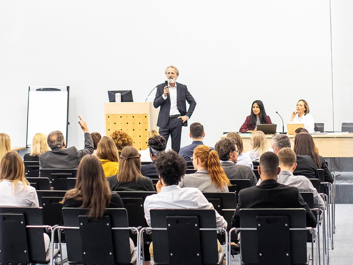 Business professional leading an in-person corporate training session in a large conference room with audience Q&amp;A.