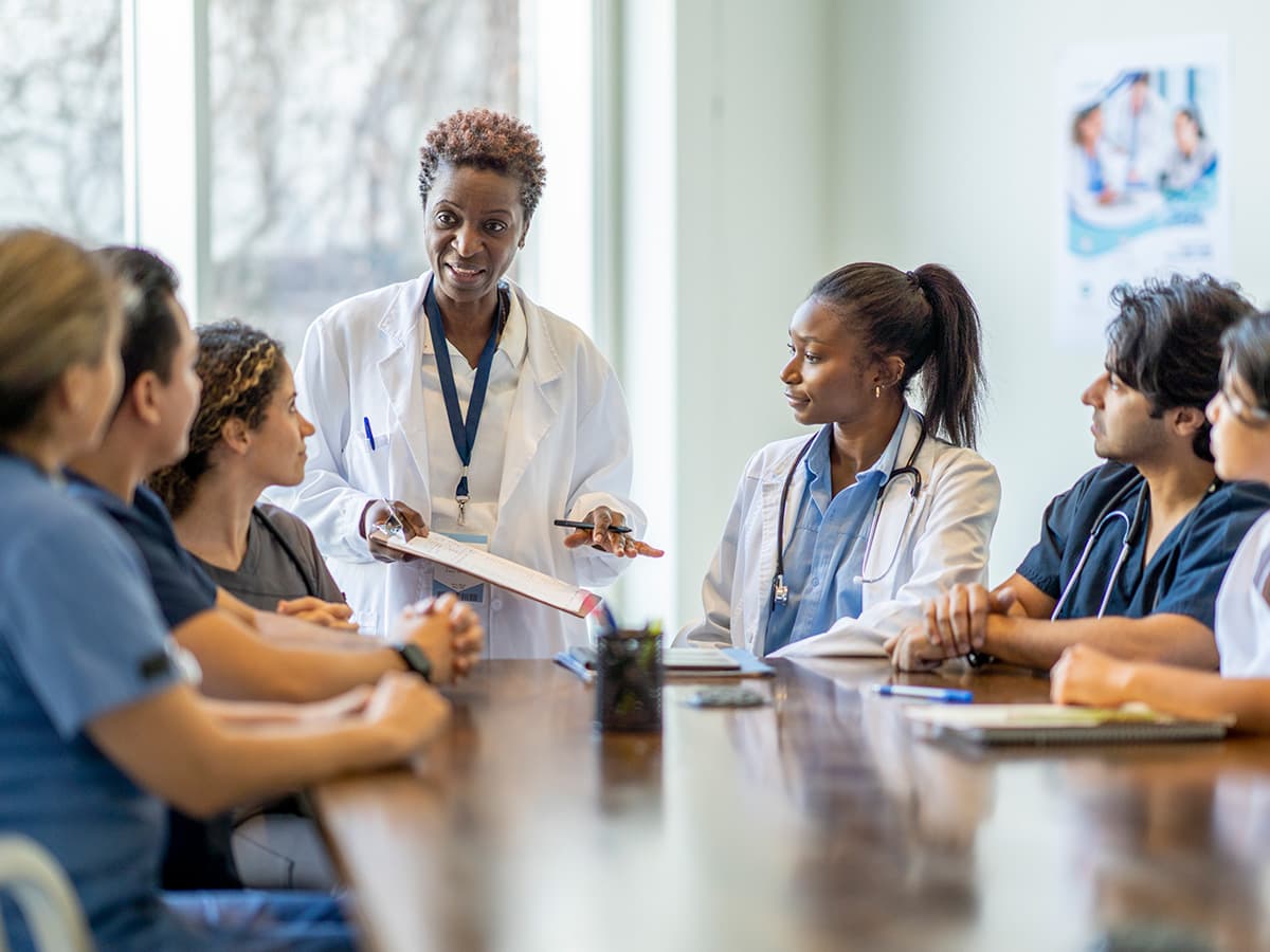 Healthcare administrator leading a team briefing with nurses and doctors around a conference table in a medical facility.