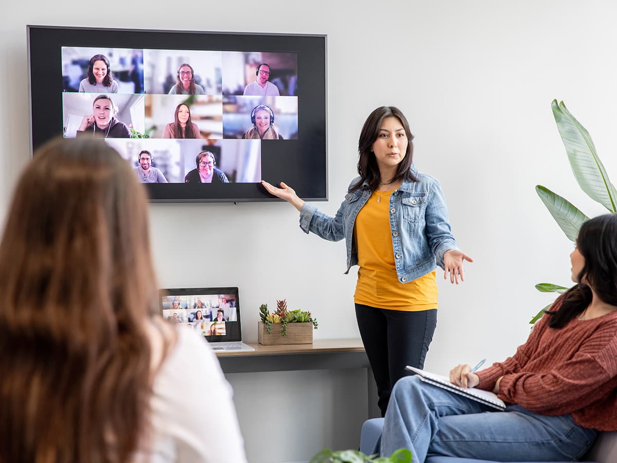 Administrator presenting to in-room and remote colleagues during a hybrid team meeting with video conferencing displayed on a wall-mounted screen.