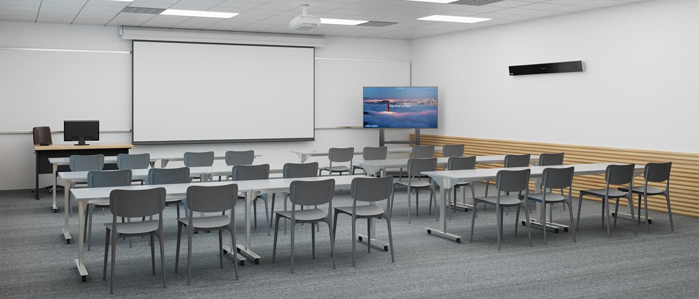 Empty standard classroom configured as a Zoom Room, featuring a Zoom-certified Nureva HDL310 audio conferencing system mounted on the wall, rows of student desks and mobile chairs, a ceiling-mounted projector with pull-down screen, and a side-mounted display showing the Zoom Rooms idle screen.