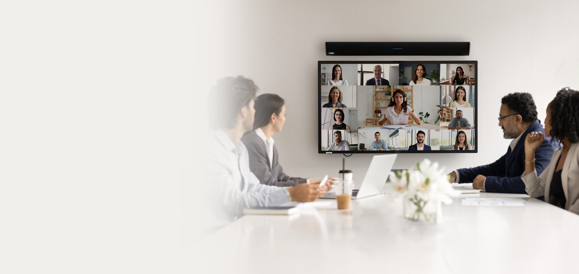 The integrated HDL310 microphone and speaker bar above a display in a meeting room, with people seated at the table and others participating remotely, shown on the display.