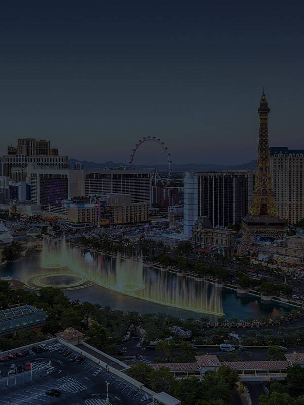 Aerial view of the Las Vegas Strip at dusk, featuring the illuminated Bellagio fountains in the foreground, the Eiffel Tower replica rising above surrounding hotels, and the High Roller observation wheel visible against the evening skyline.