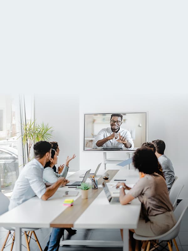 Diverse business team seated around a conference table collaborating with a remote colleague displayed on a wall-mounted screen during a hybrid meeting.
