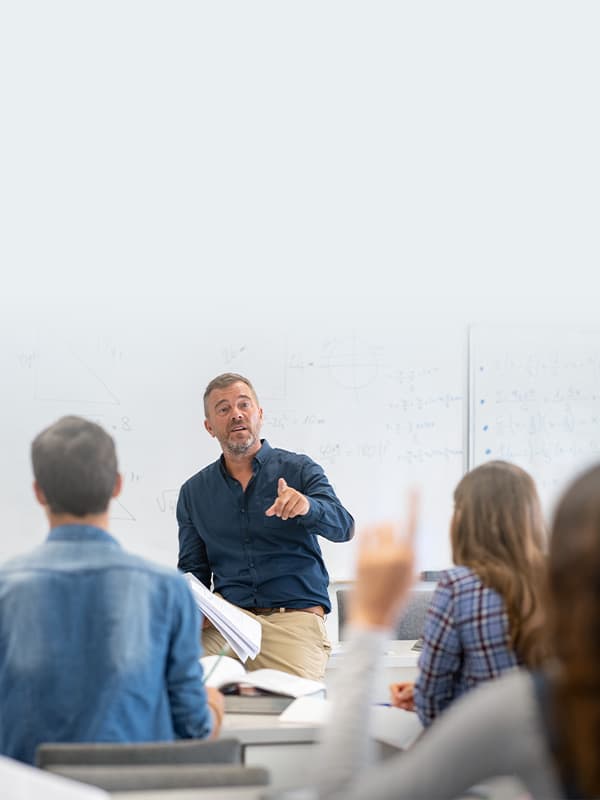 University instructor engaging with students during an in-person classroom session, with a student raising their hand to ask a question in front of a whiteboard.