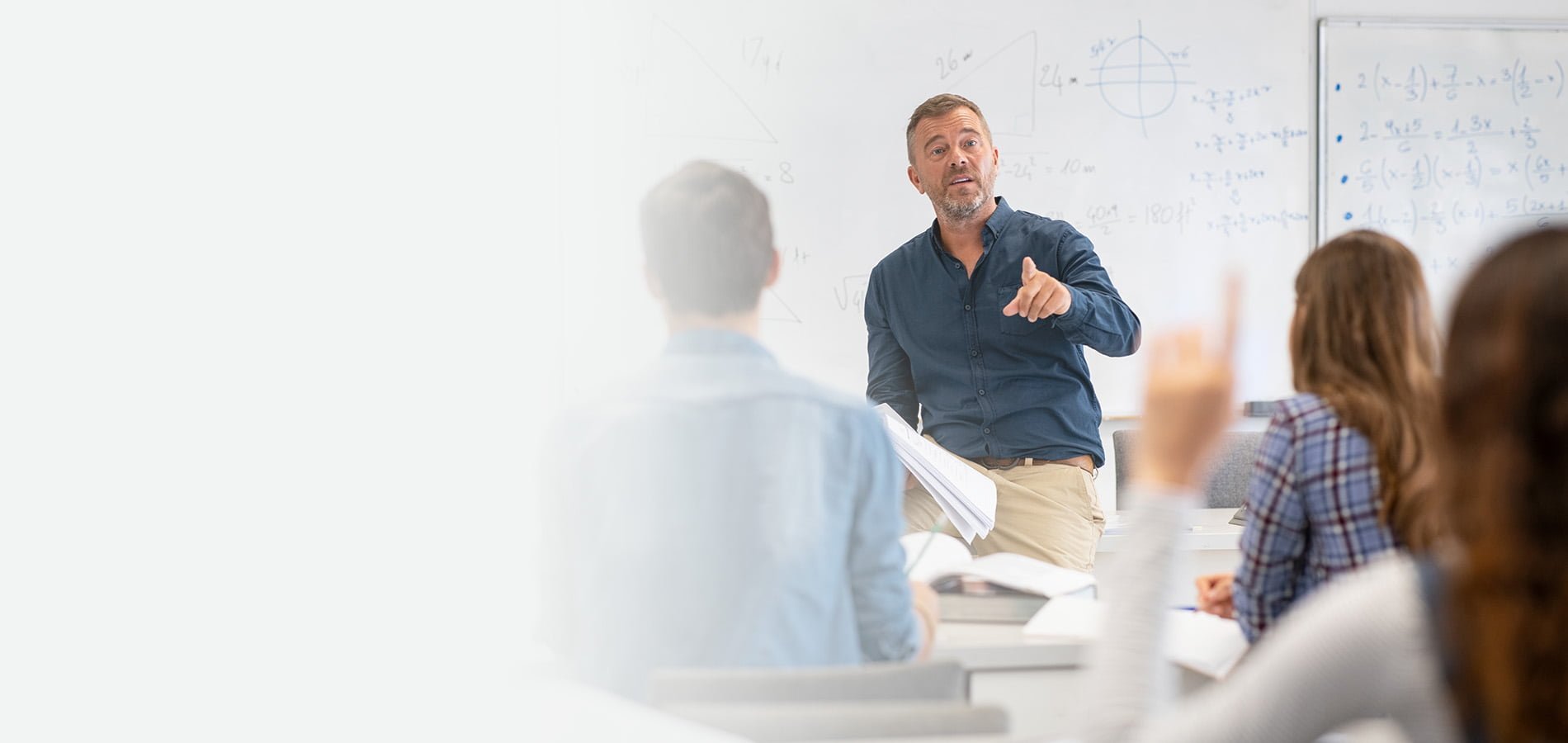 University instructor engaging with students during an in-person classroom session, with a student raising their hand to ask a question in front of a whiteboard.