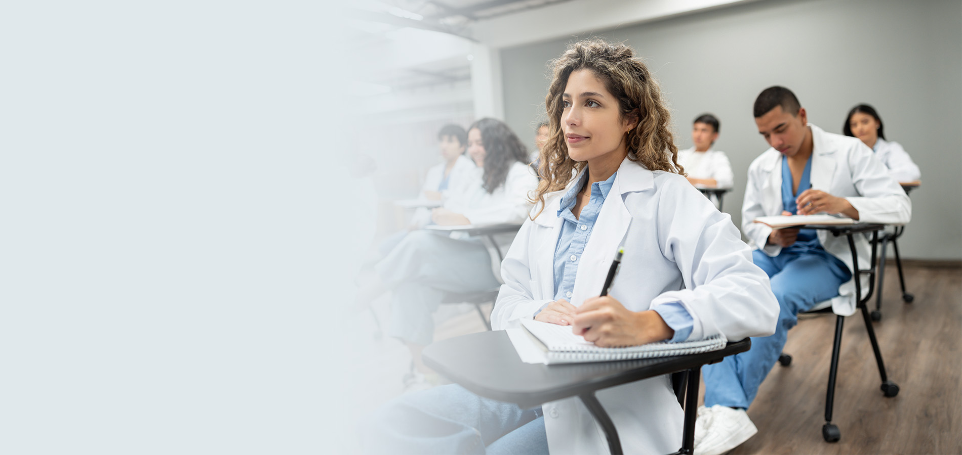 Medical students in white coats taking notes attentively during a healthcare training session in a classroom setting.